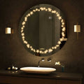 A round bathroom mirror with LED lights around the circumference. The mirror is mounted on a bathroom wall above a sink. There is text written on the bottom right corner of the mirror that says Glazonoid.
